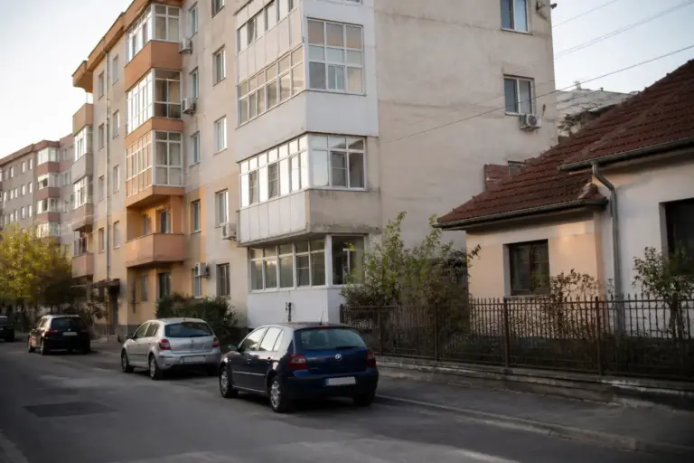 O stradă cu o casă veche în prim-plan și un bloc de locuințe cu mai multe etaje în fundal, cu mașini parcate pe marginea drumului sub lumina soarelui la apus