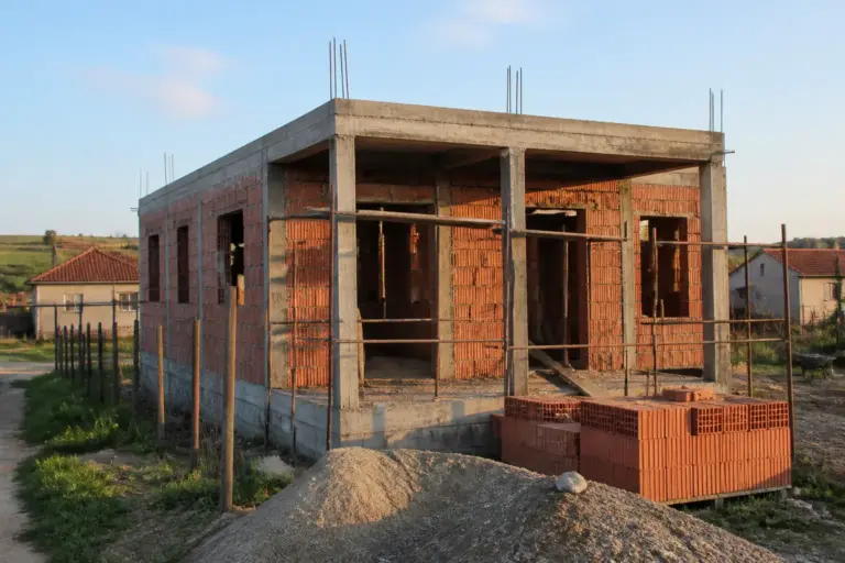 O casă aflată în stadiul de construcție la roșu, realizată din cărămidă și beton, situată într-o zonă rurală cu alte case vizibile în fundal